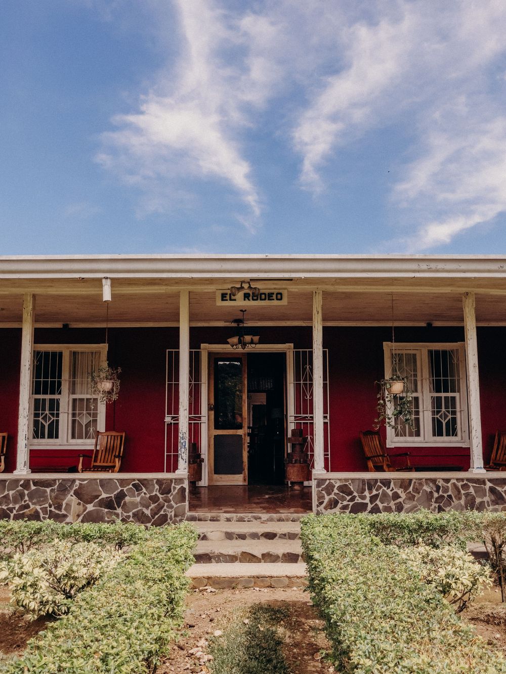 Red ranch-style building with stone base, white porch, and El Rodeo sign under a blue sky.