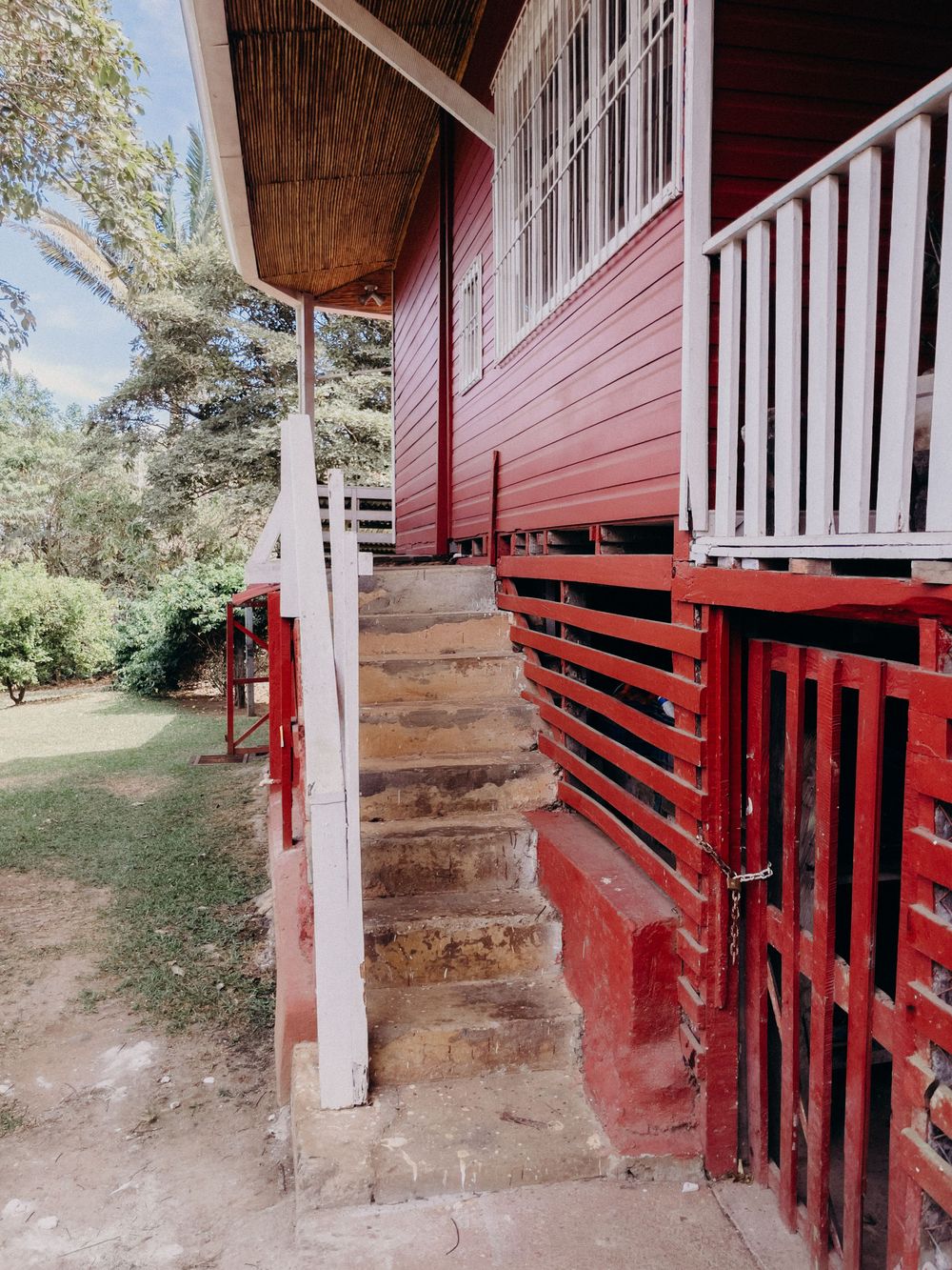 Red wooden house exterior with stone steps, white railings, and a fenced porch surrounded by lush green trees.