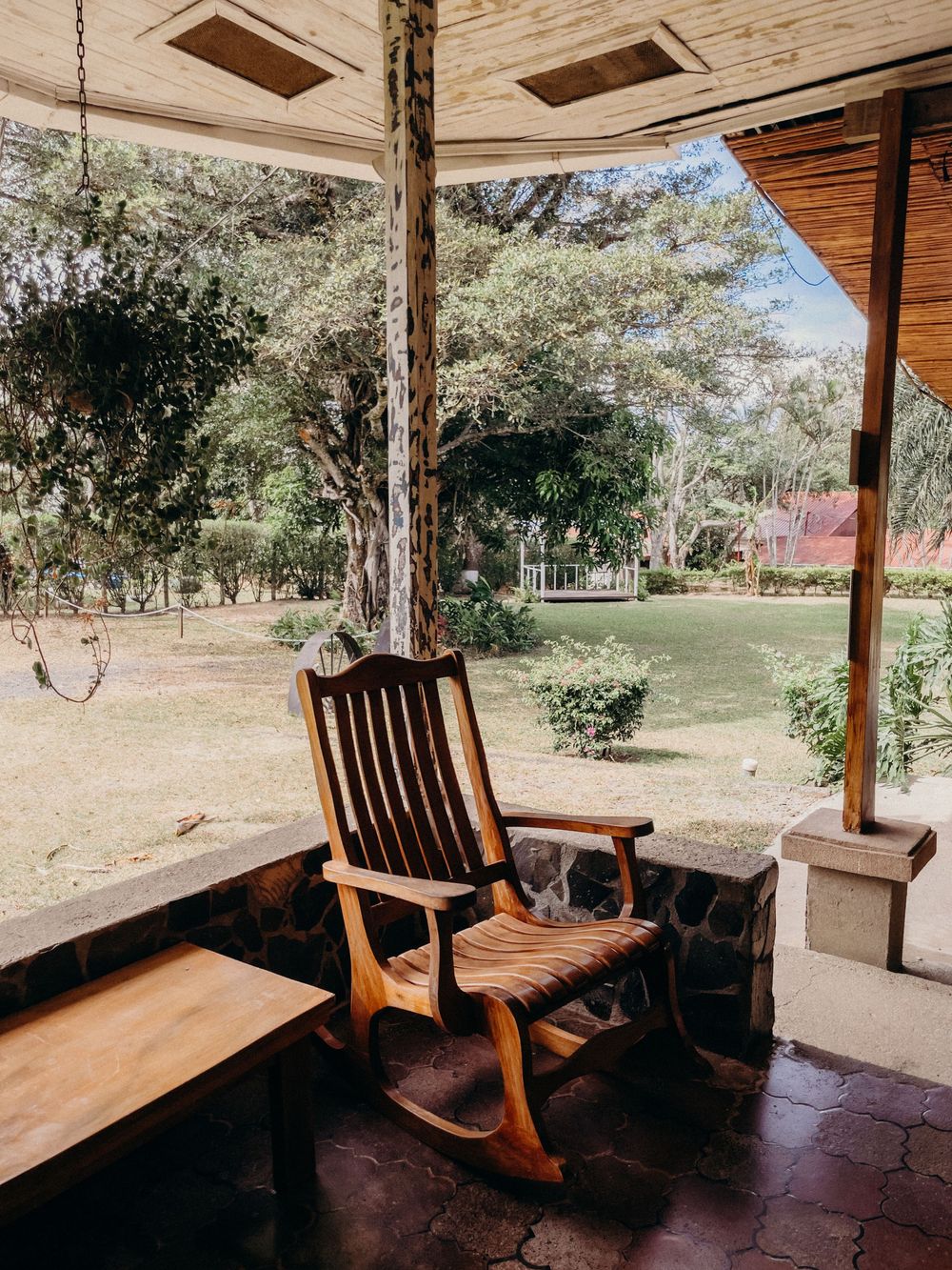 Wooden rocking chair on a rustic stone porch overlooking a lush green garden with tall trees.