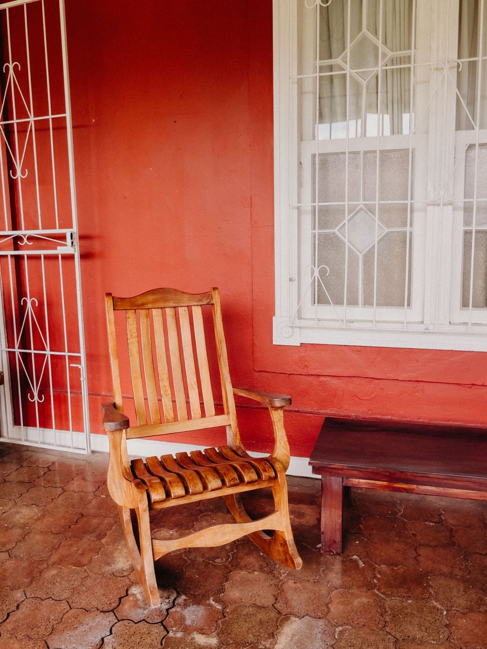 Wooden rocking chair on a red porch beside a bench, with barred windows and tiled flooring.