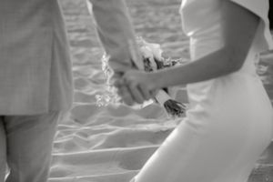 Black and white photo of bride and groom holding hands and bouquet on sandy beach at their wedding.