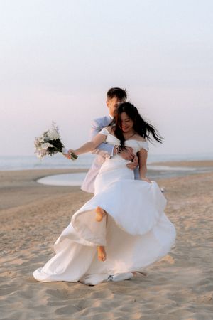 A joyful couple celebrates their beach wedding, with the groom lifting the bride in her flowing white gown as she holds a bouquet.