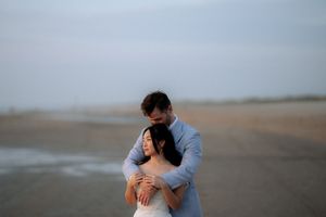 Couple embracing tenderly on a misty beach, man in blue shirt hugging woman in white dress near the ocean shoreline.