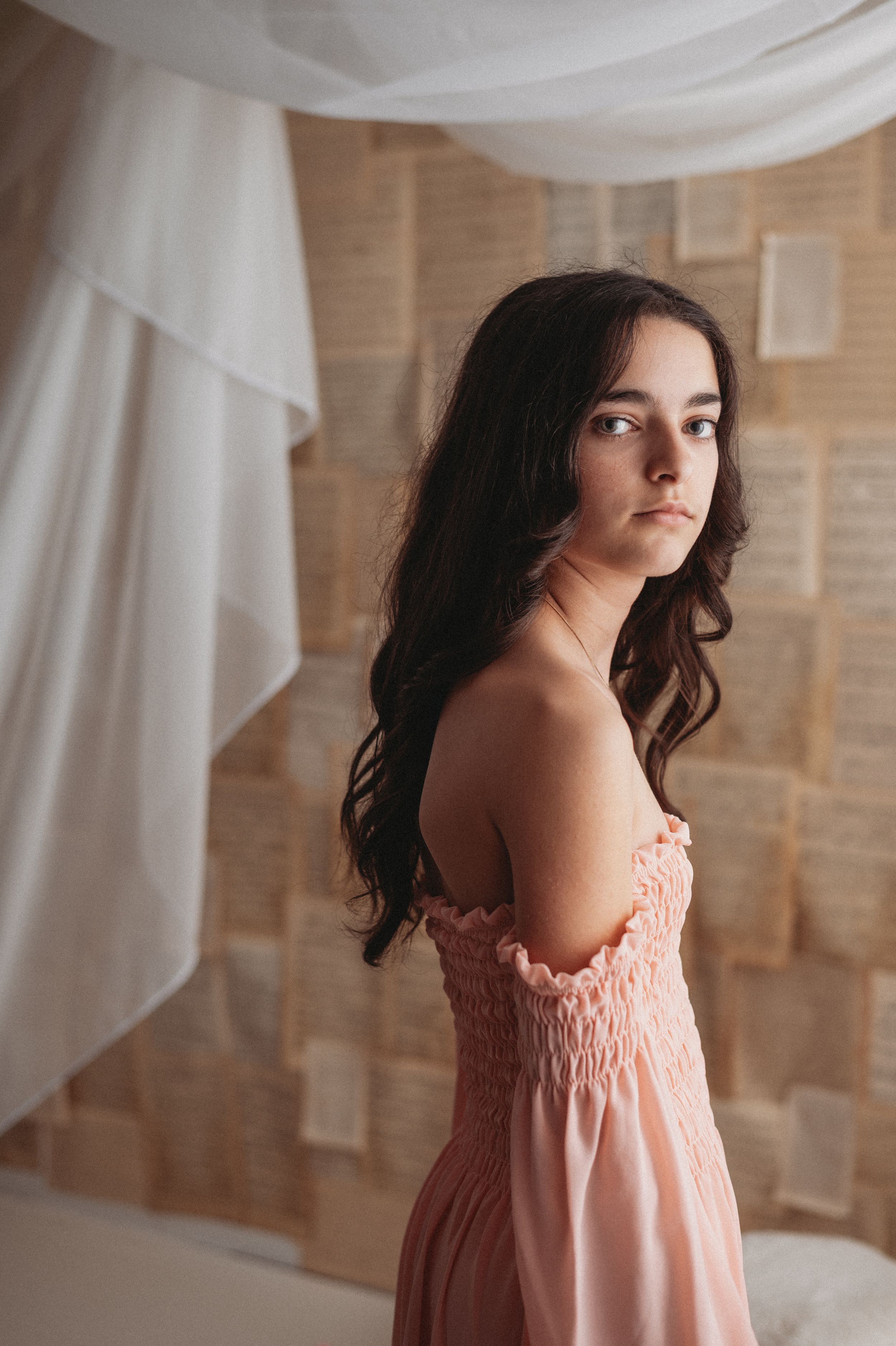 Soft romantic portrait in pink off-shoulder dress against white curtains and wooden wall background.