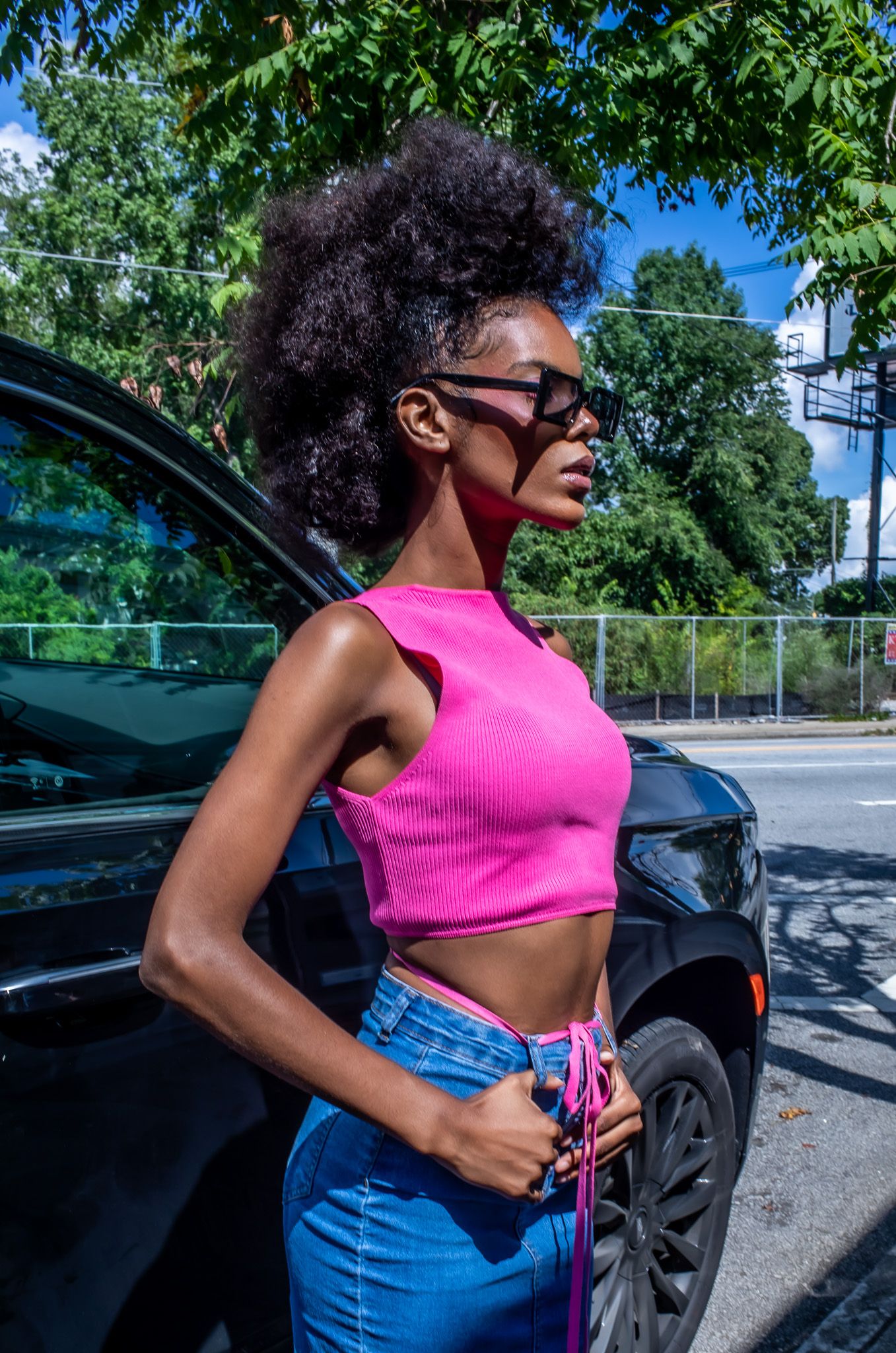 Person in pink crop top and jeans poses against a black car on a sunny day.