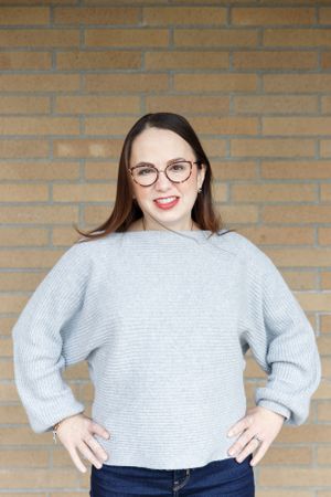 Portrait of someone in a light gray sweater and glasses against a brick wall.