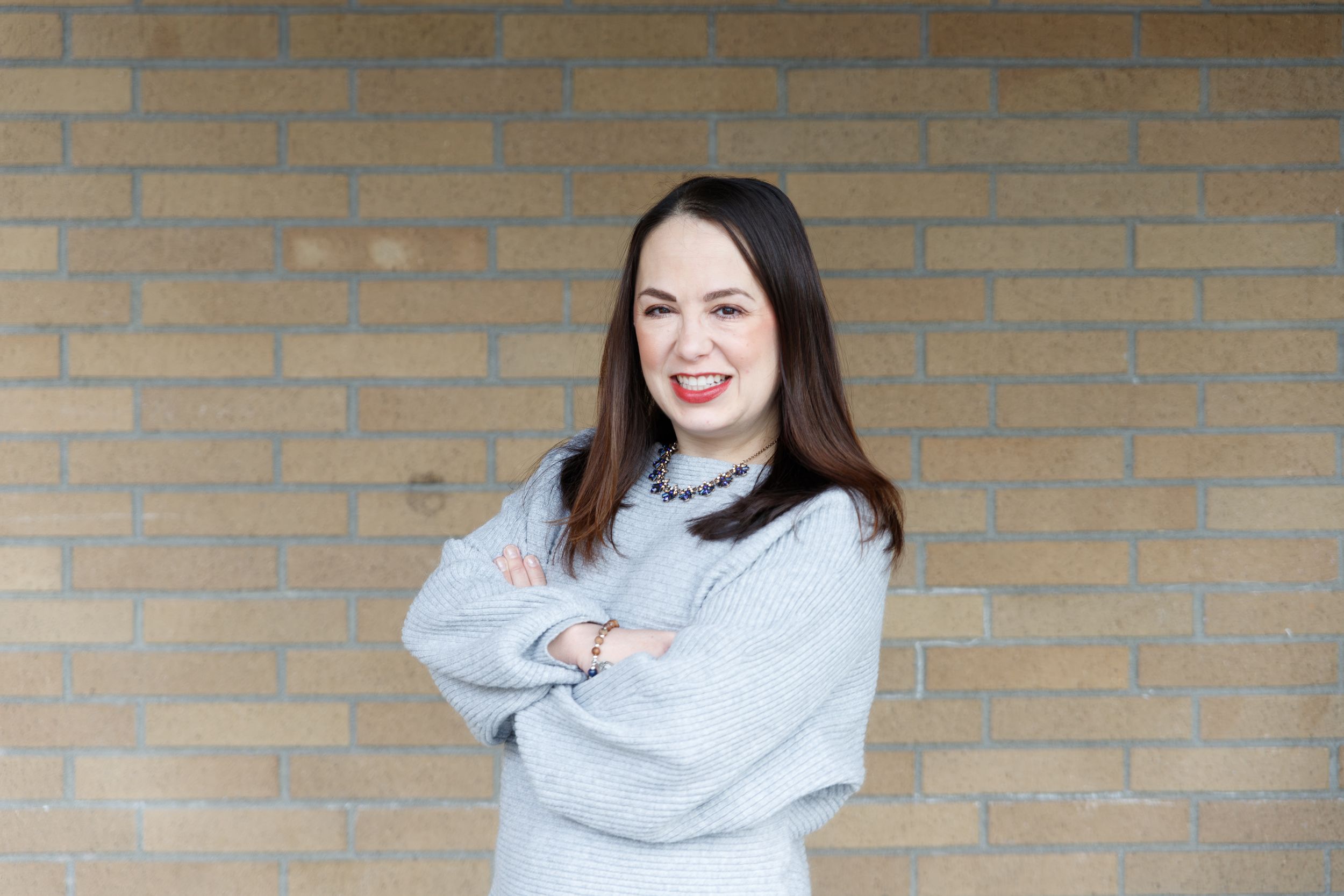 Professional portrait of person in light gray sweater against brick wall background.