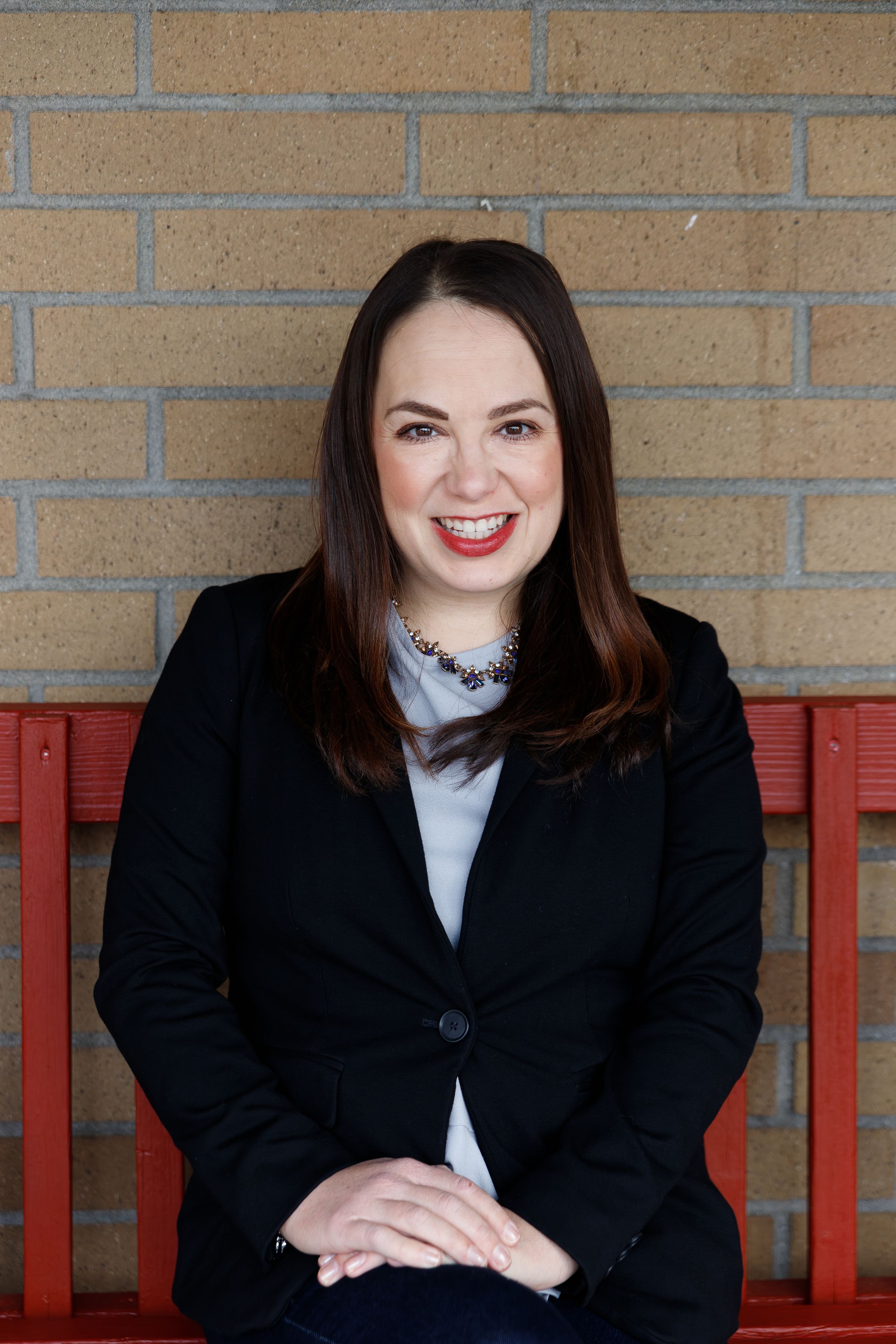 A professional headshot of someone in business attire sitting on a red bench against a brick wall.