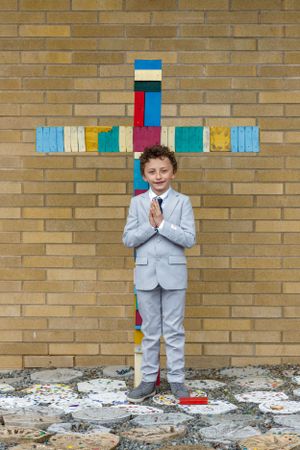 Child in gray suit standing in front of colorful block cross on brick wall.