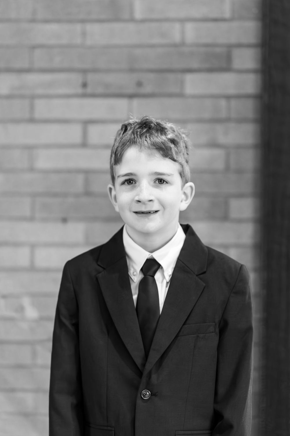 Formal portrait of young person in black suit and tie against brick wall.