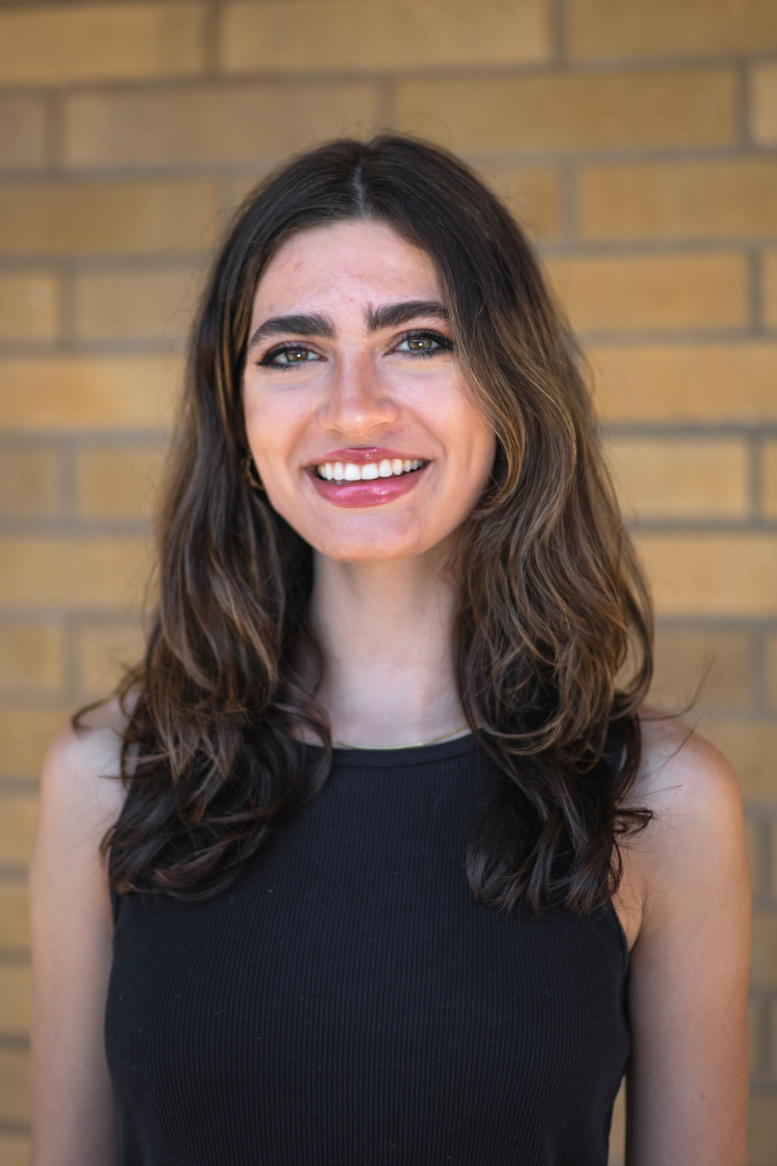Professional headshot of smiling person in black sleeveless top against yellow brick wall.