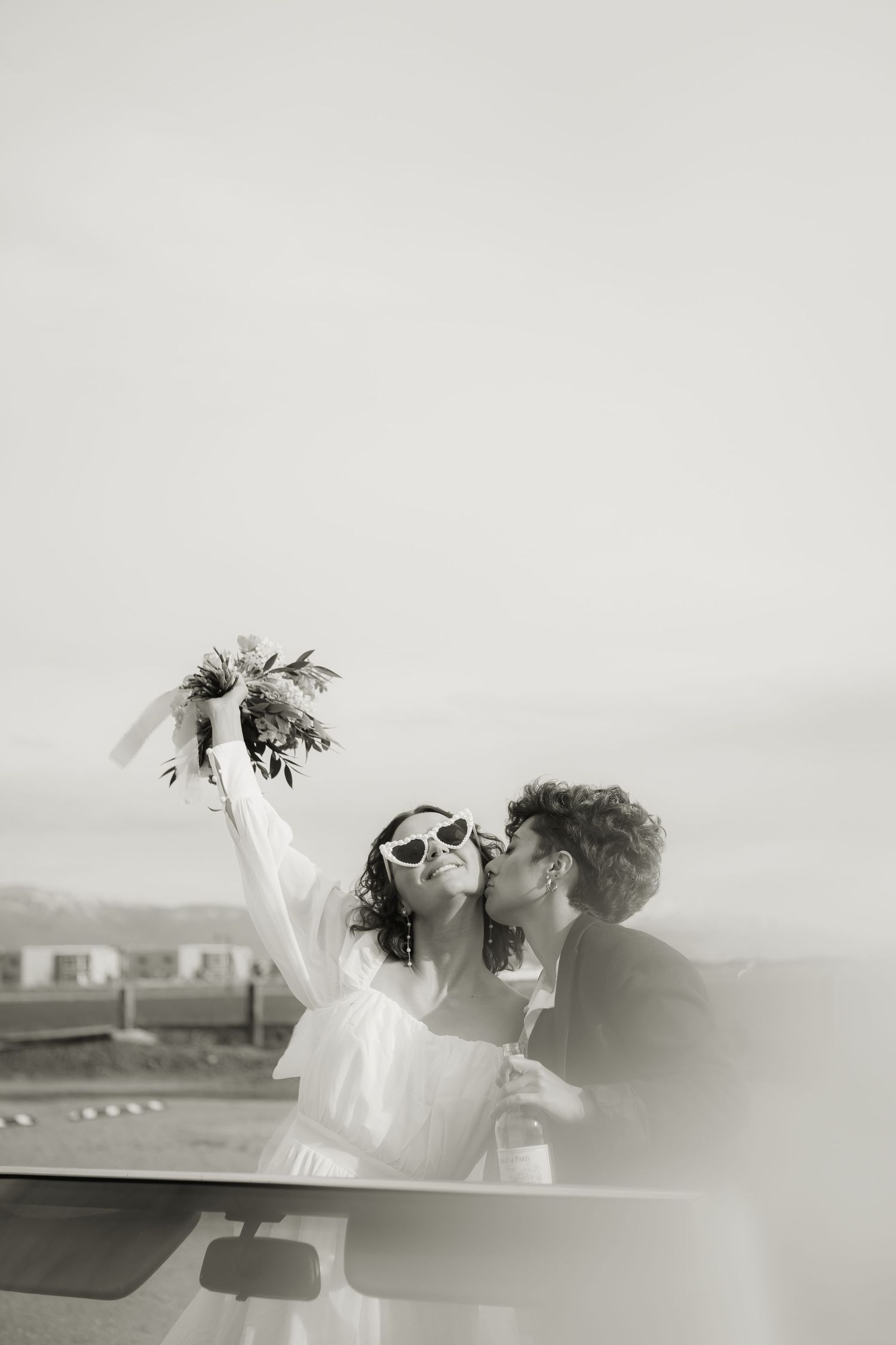 Black and white wedding photo of couple kissing next to car with bouquet.