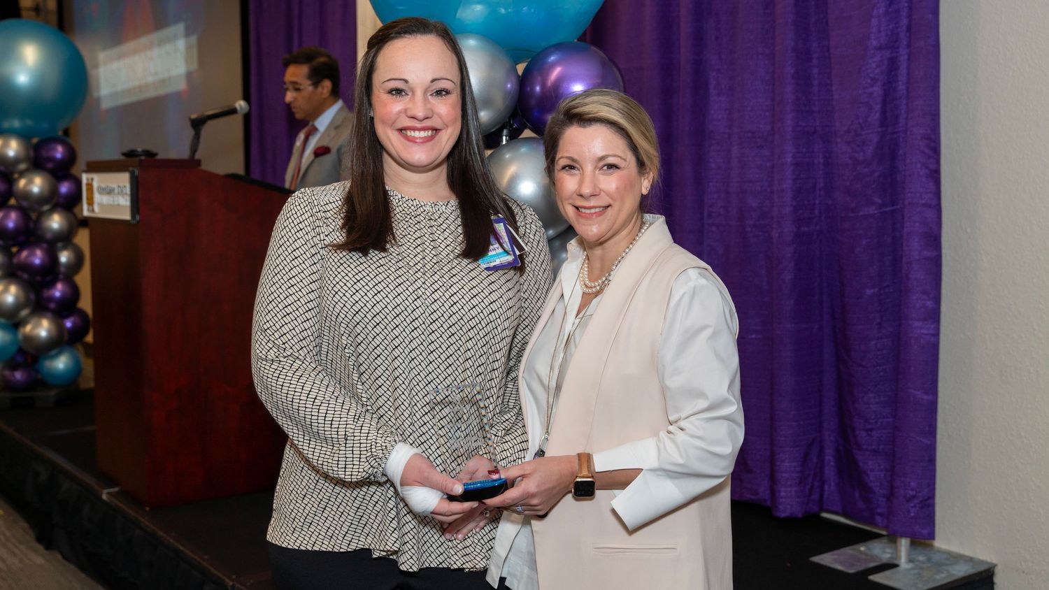 Two women pose with an award at a formal event with purple backdrop.