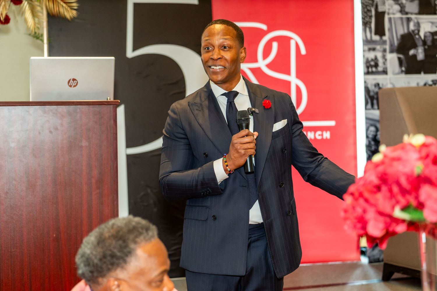A speaker in a navy suit holds a microphone while presenting at a podium with a red backdrop and flowers nearby.