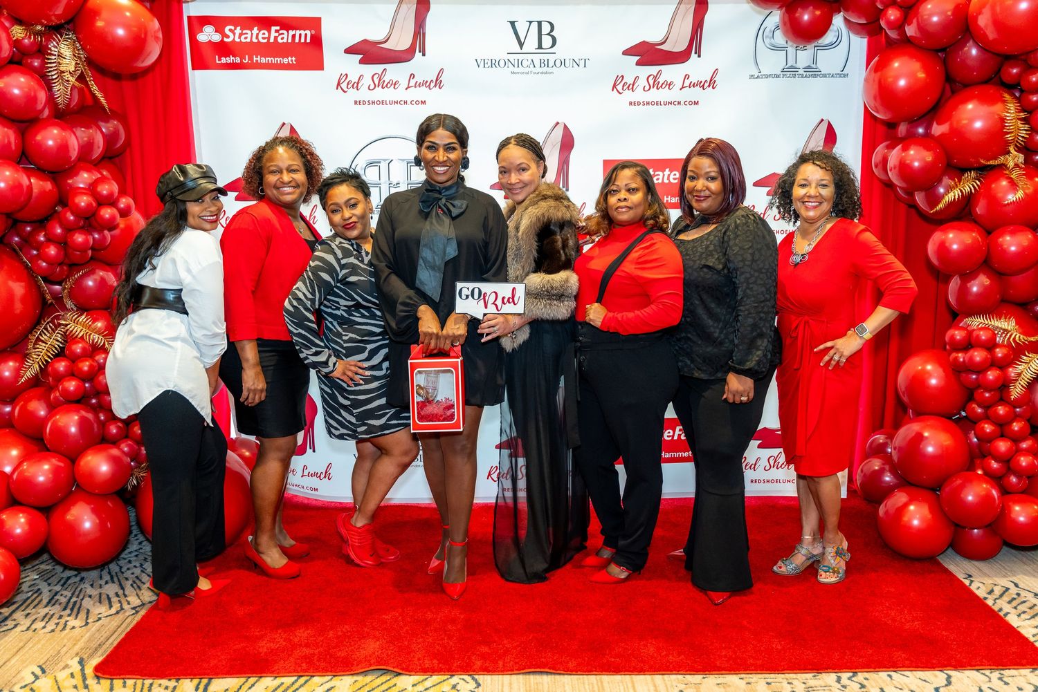 A group poses on a red carpet at an event decorated with red balloons and sponsor banners in the background.