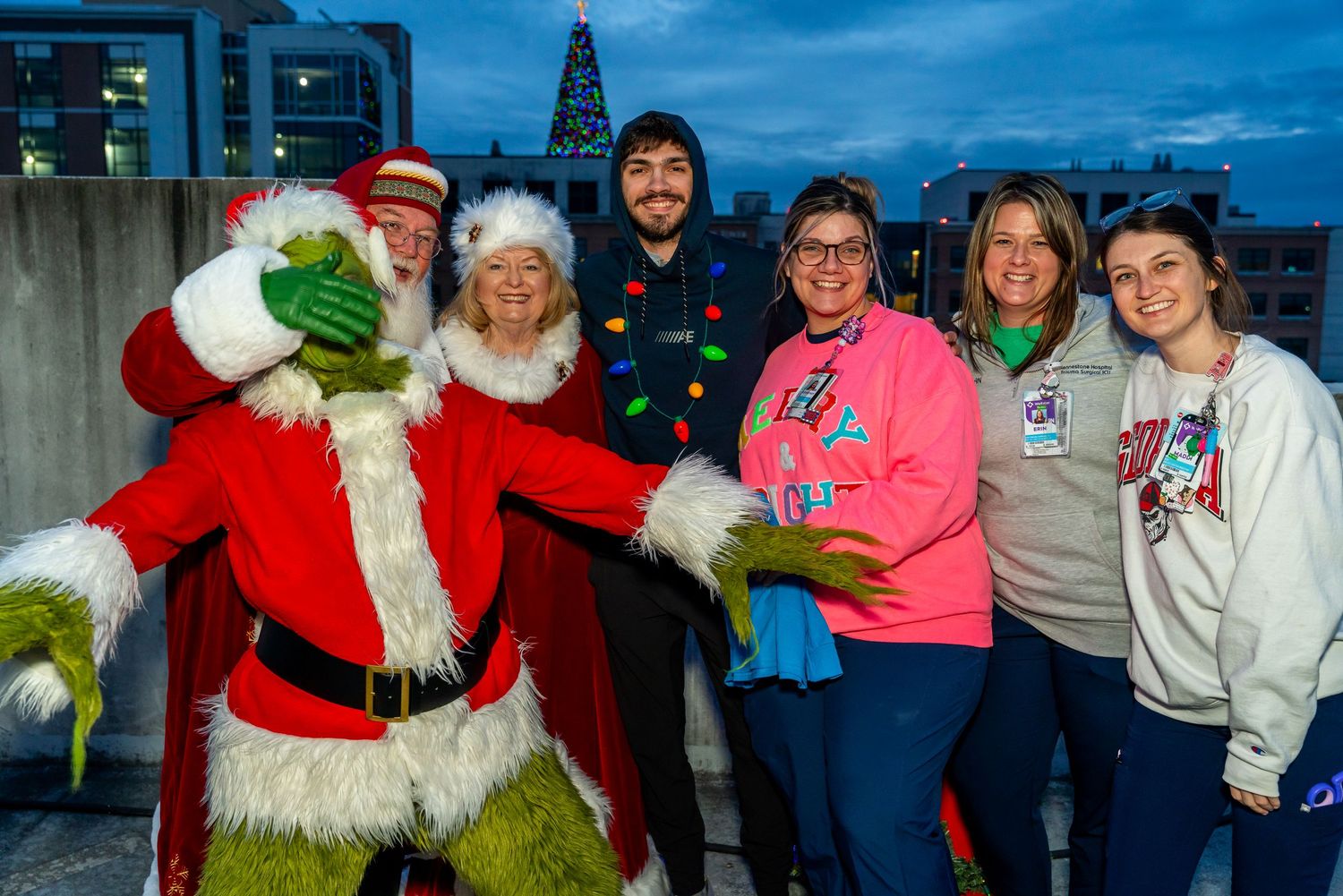 A group wearing festive holiday attire poses with the Grinch in a Santa suit during an evening outdoor event.