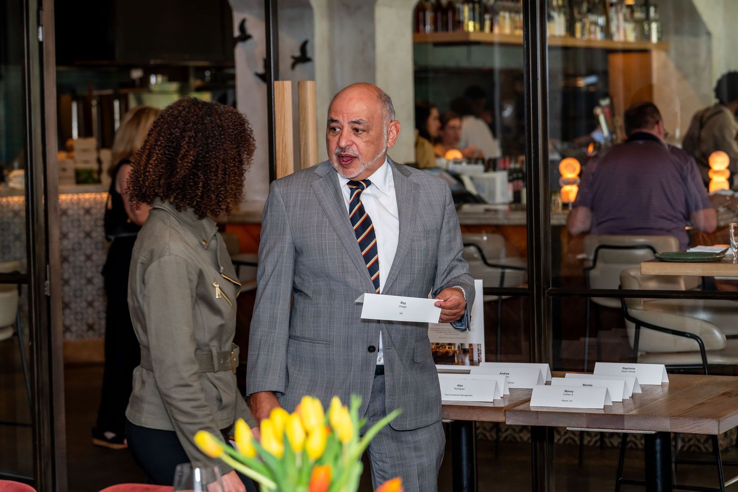 A restaurant server in a gray suit and striped tie delivers plates at a dining establishment with yellow tulips in view.