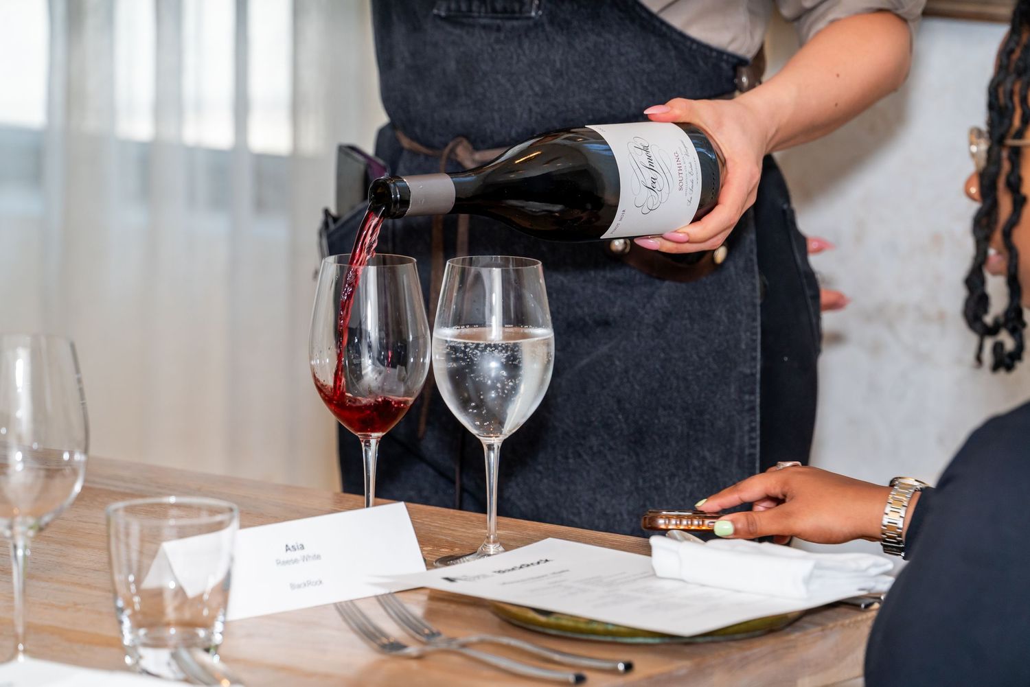 A server wearing a dark apron pours red wine into glasses at a table set with menus and glassware.