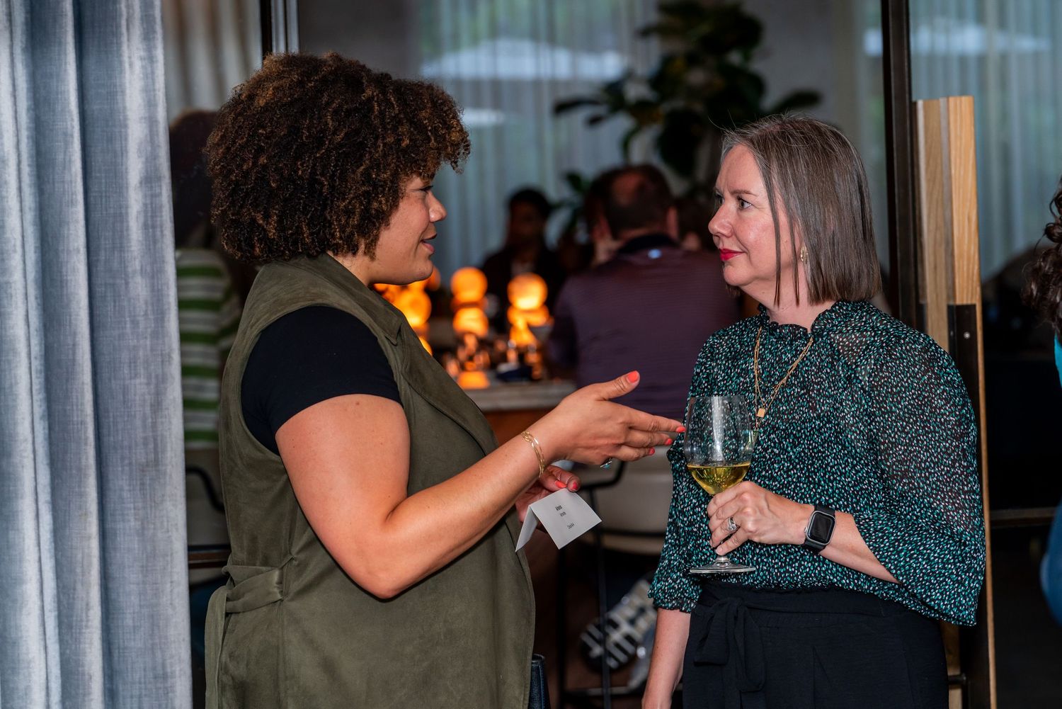 Two people in deep conversation while holding drinks at an evening gathering with warm lighting in the background.