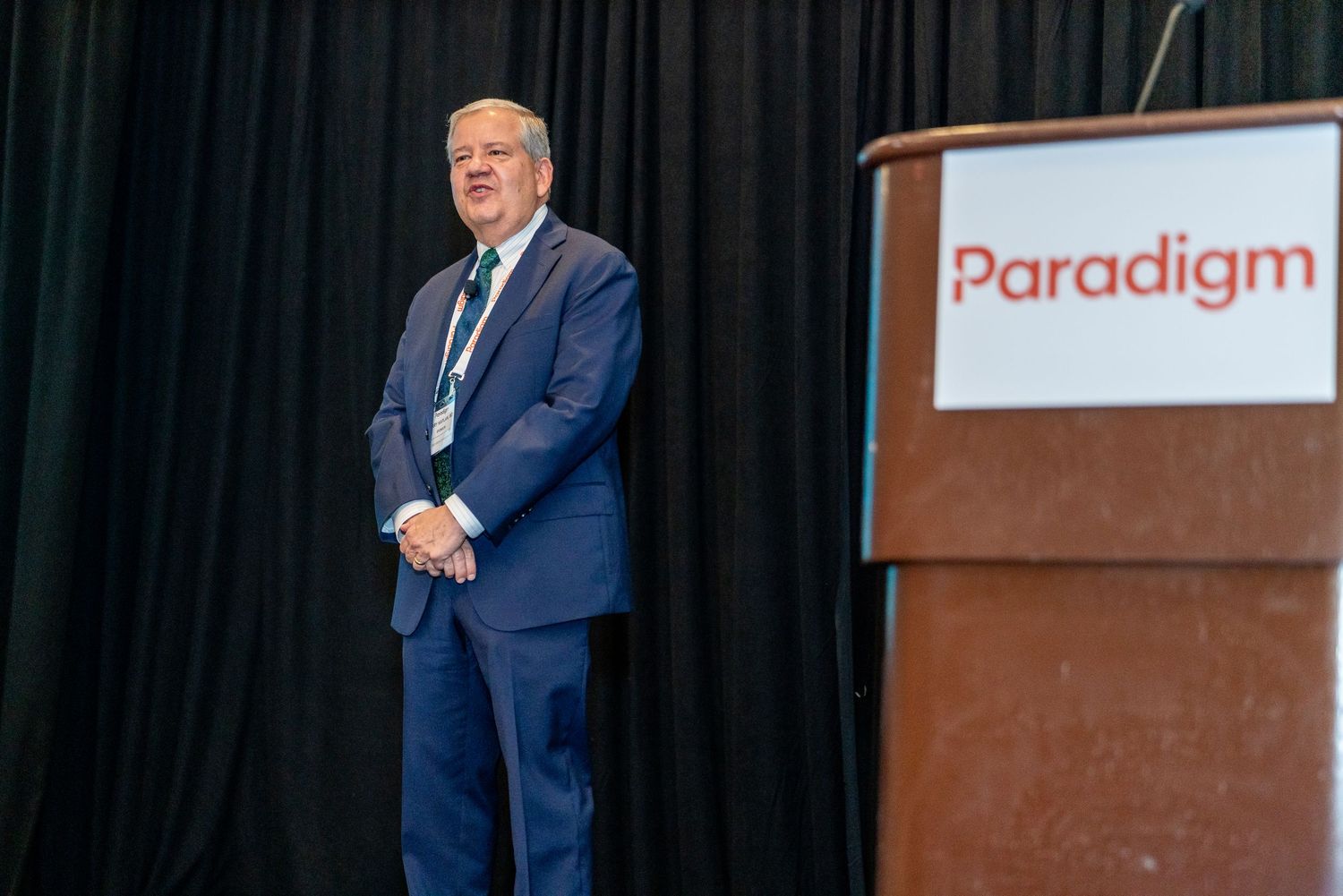 Speaker presents on stage at a corporate event with 'Paradigm' branding visible in the background.