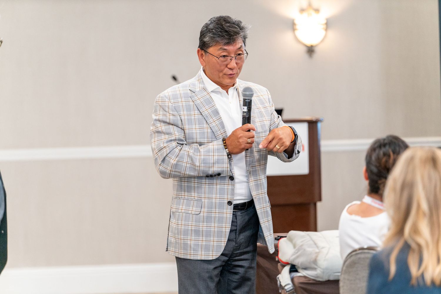 A speaker in business attire delivers a presentation while gesturing to an audience in a bright conference room setting.