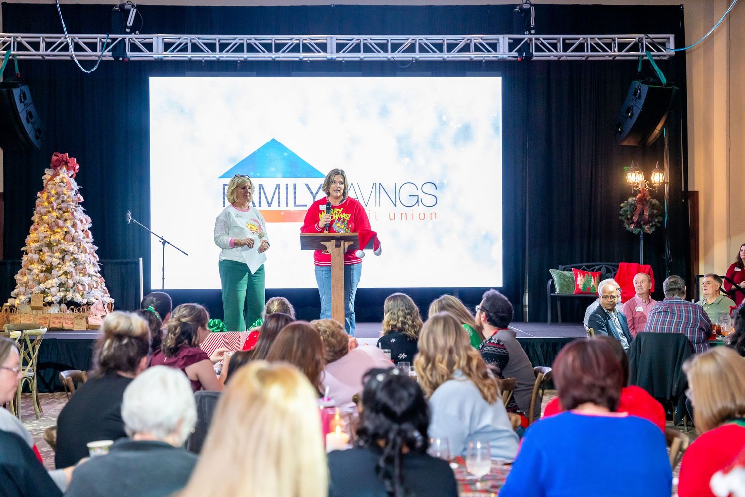 A festive holiday event with speakers on stage addressing an audience near a decorated Christmas tree.