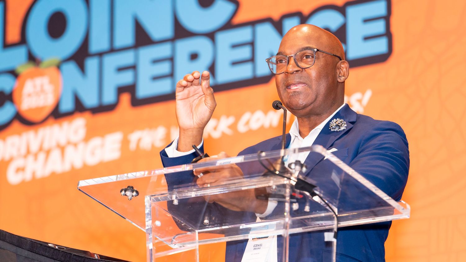Speaker delivers passionate keynote address at conference from clear podium with orange background.