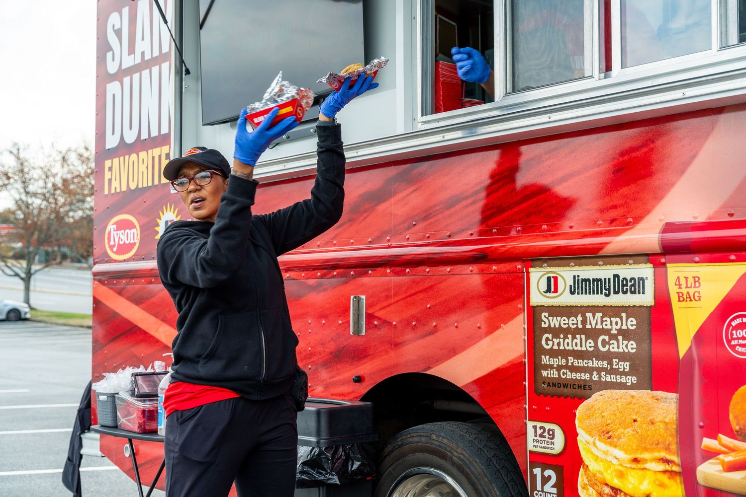 A worker stands near a bright red food truck raising up bread loaves while wearing a dark jacket and cap.