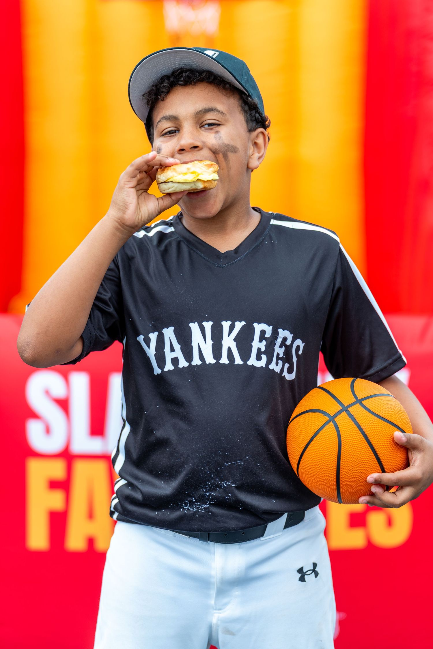 Young athlete in Yankees jersey takes a bite of food while holding basketball against vibrant red background.
