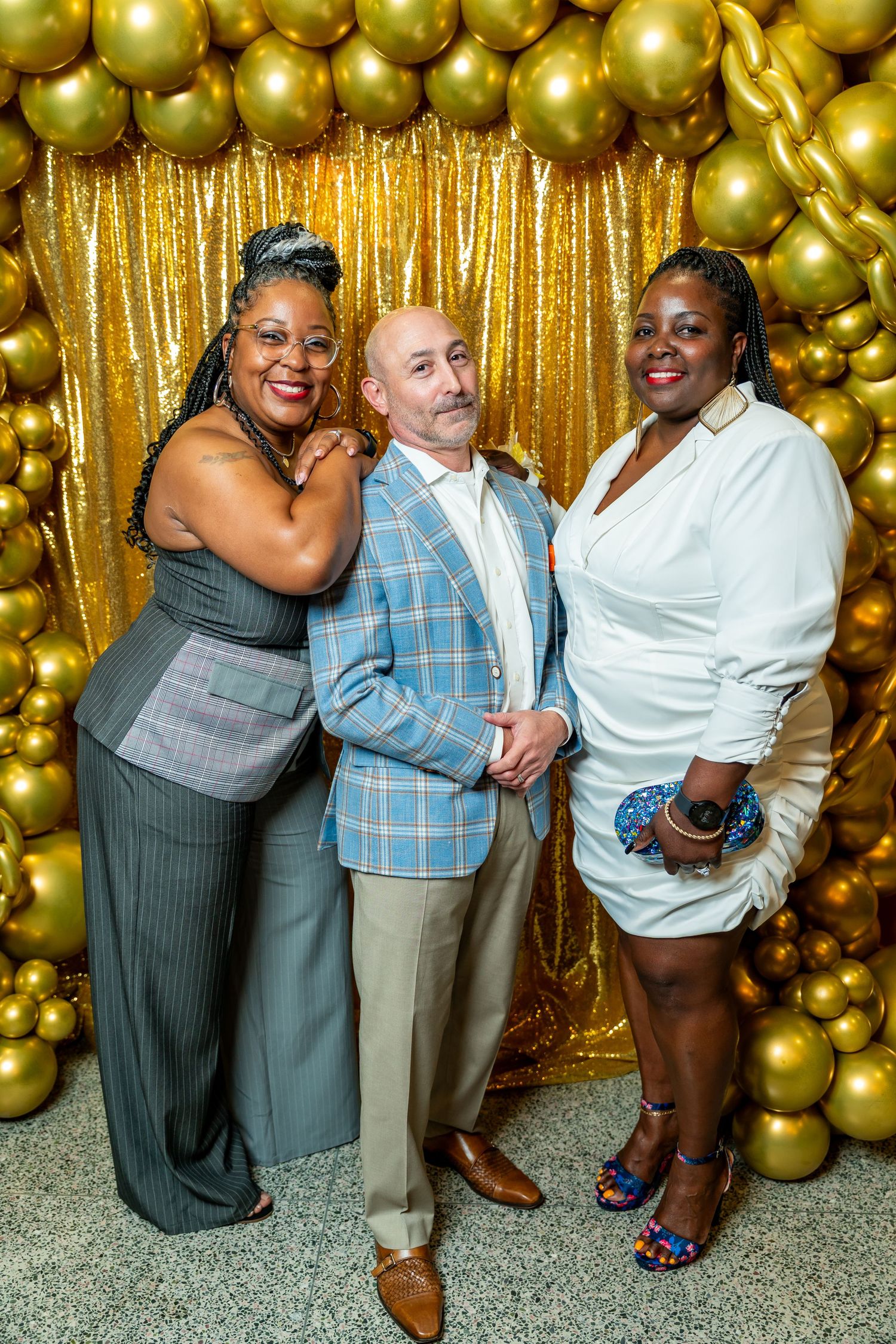 Three people pose together in front of a gold balloon wall and sparkly curtain backdrop at a festive celebration.