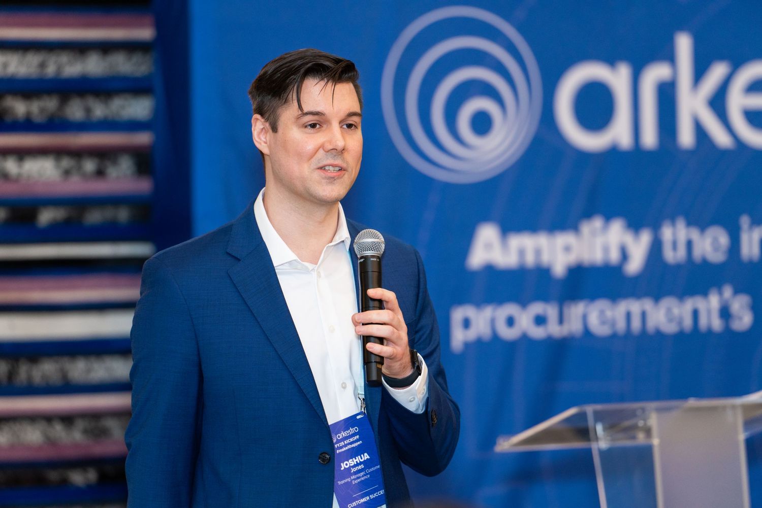A speaker in a blue suit presents at a corporate event with Arkea branding in the background.
