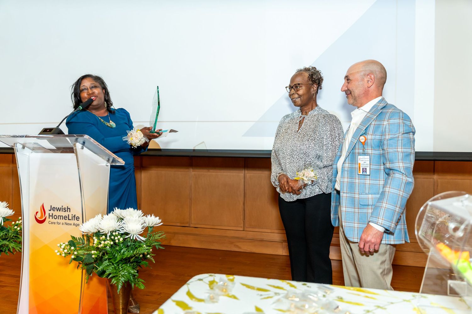 Three people interact at a podium during a professional event, with greenery decorating the presentation area.