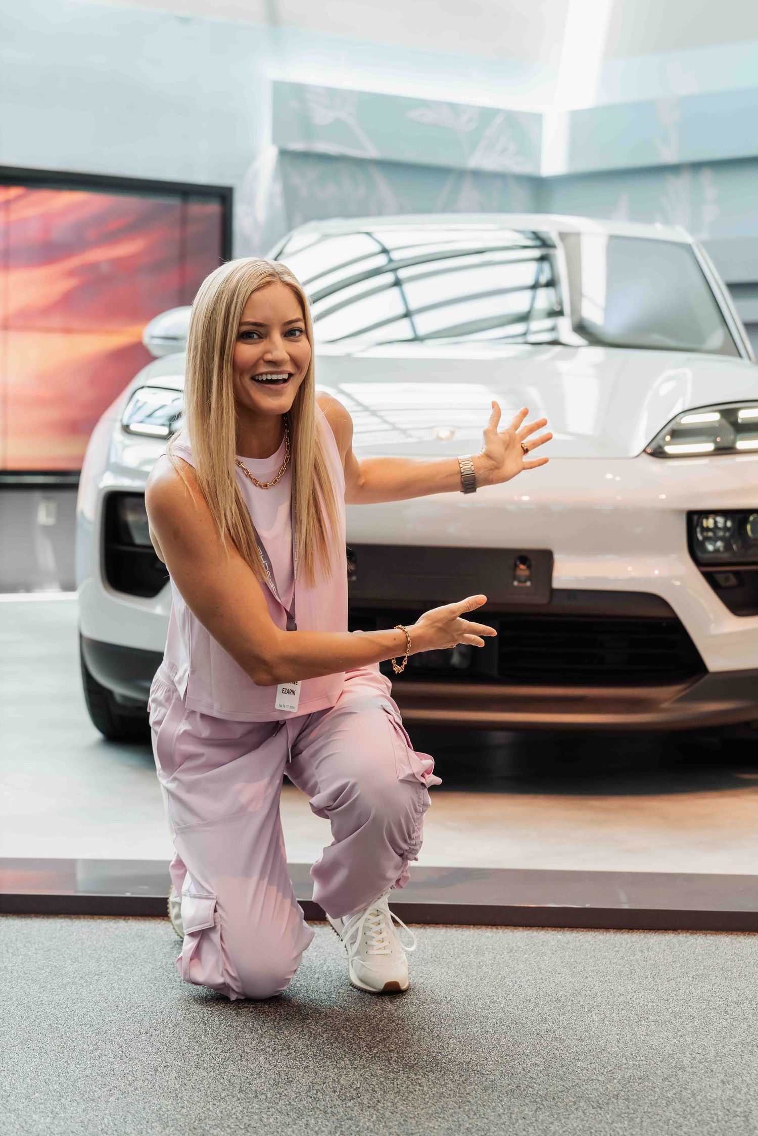 Enthusiastic person in pink outfit gestures excitedly in front of a silver luxury car.