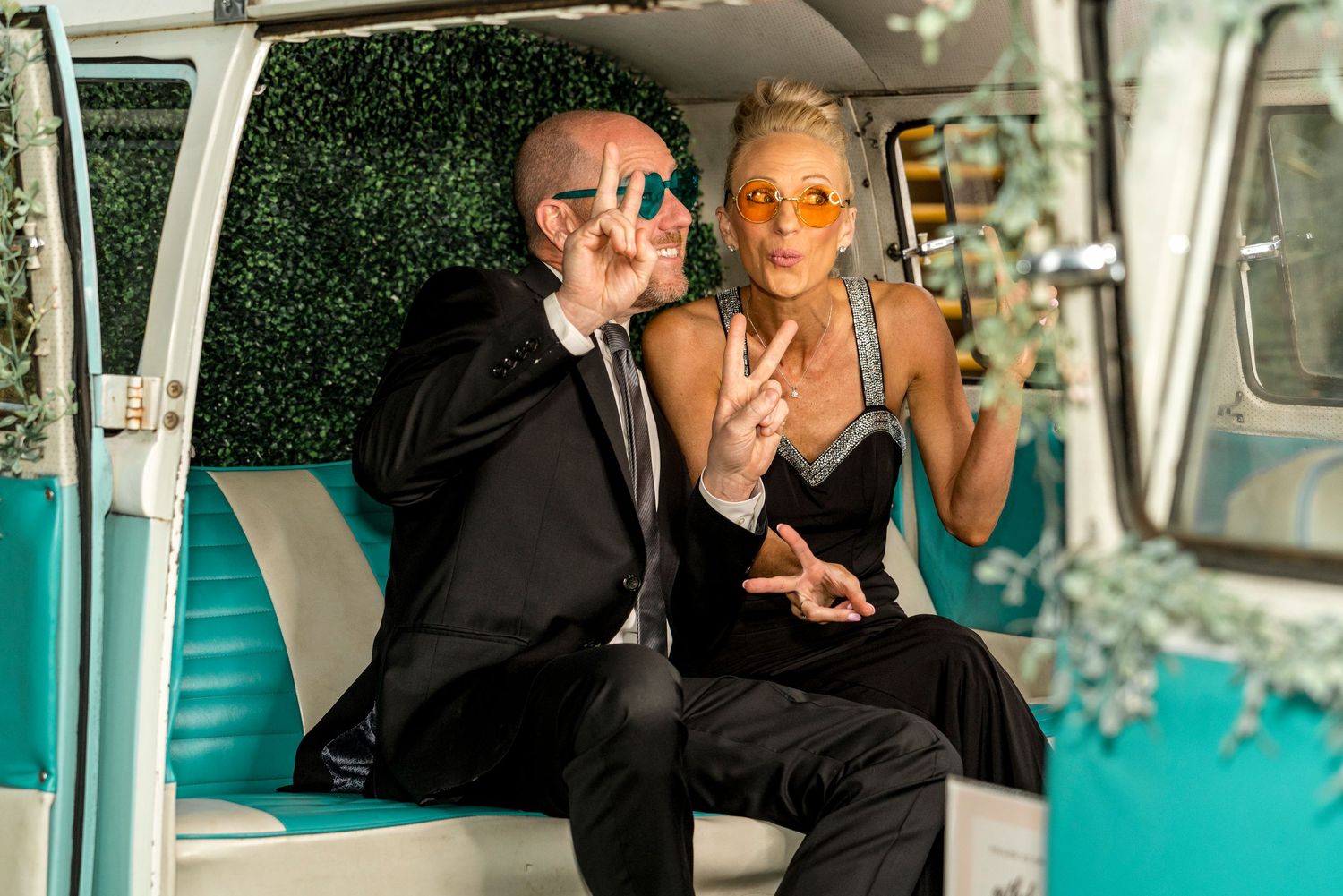 Couple in formal attire making peace signs while sitting in vintage turquoise vehicle.