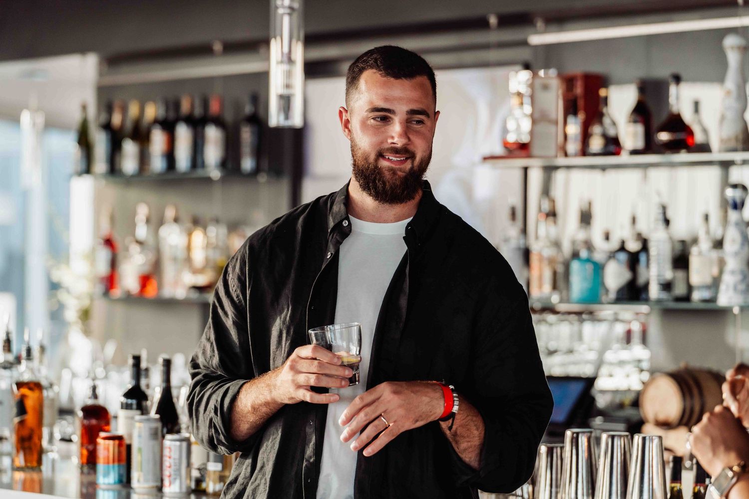 Bartender in casual attire holding a drink behind a well-stocked bar counter.