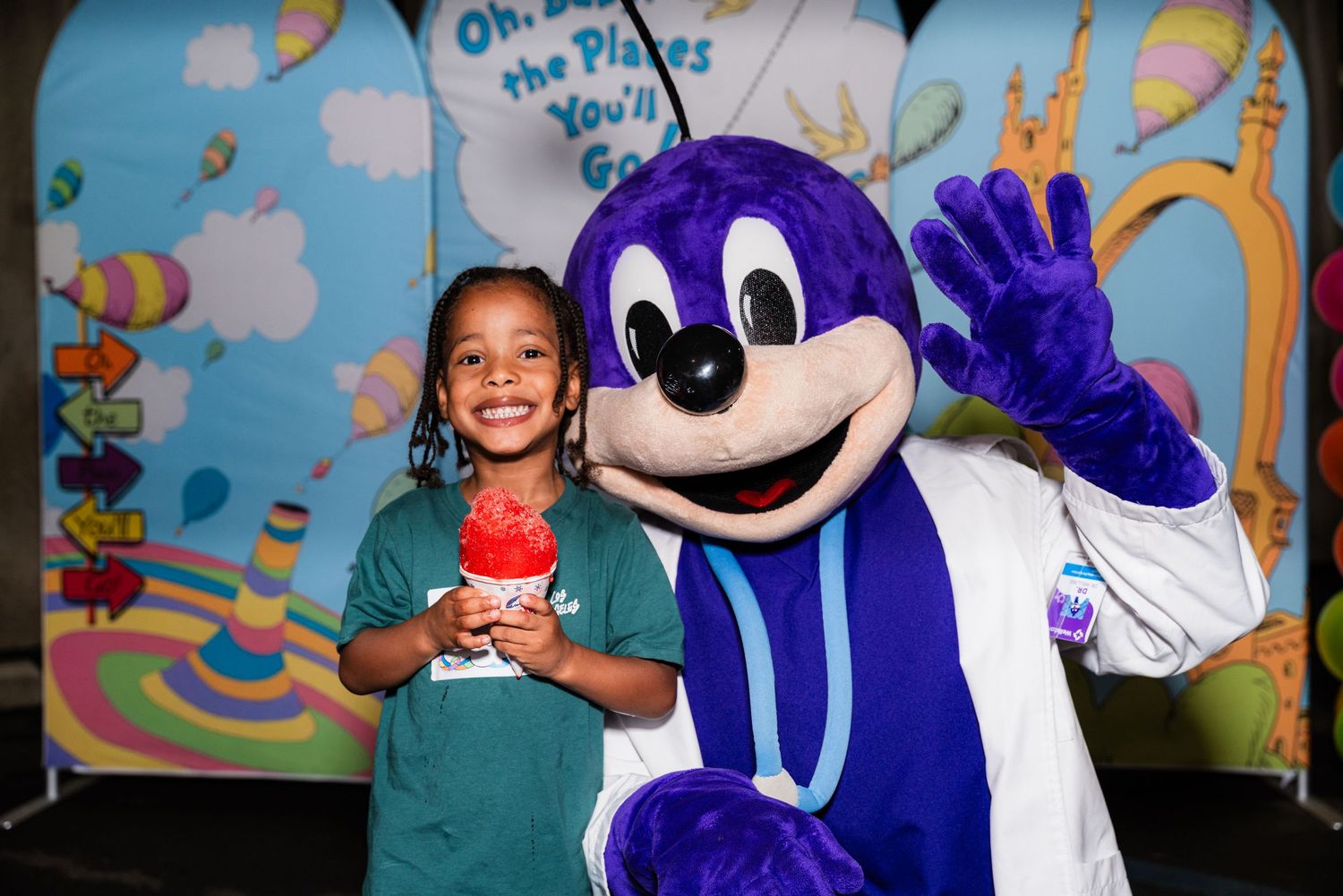 Child in teal shirt poses with purple mascot character against cartoon backdrop.