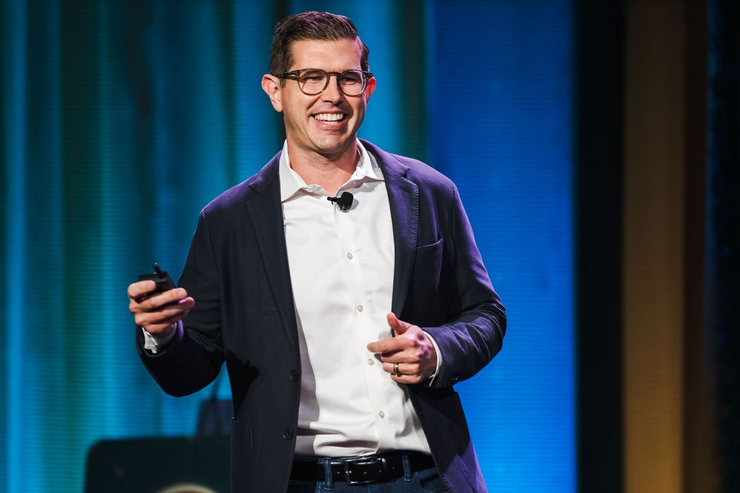 A speaker in a dark blazer and glasses presents on stage holding a mobile device against a blue backdrop.