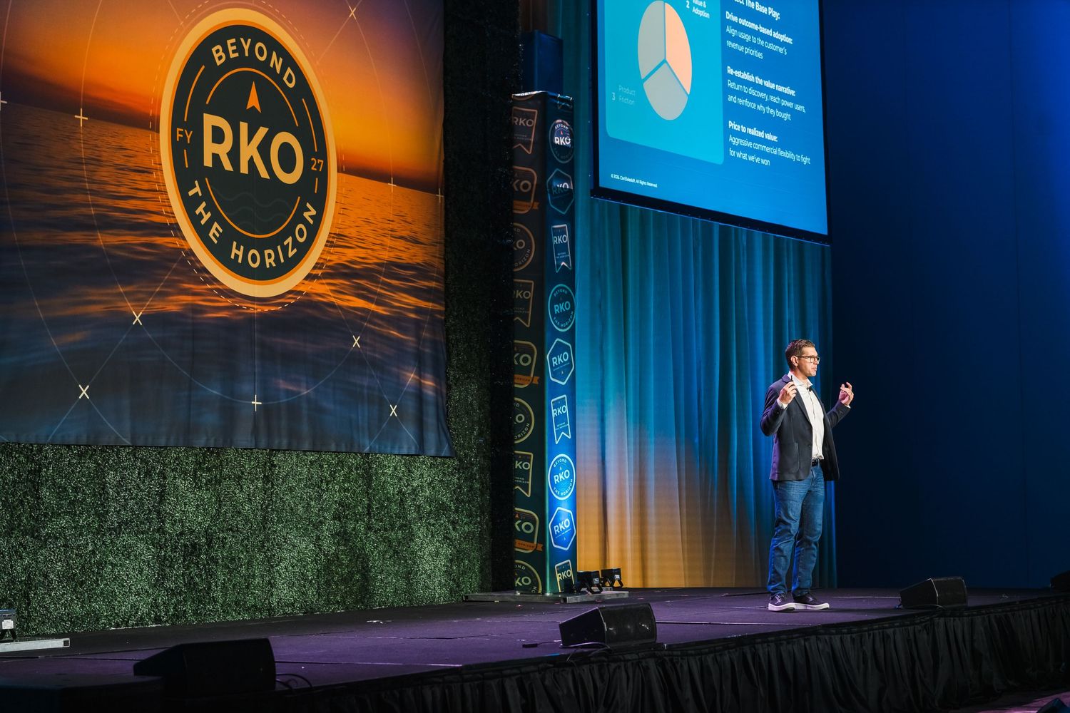 A speaker presents on stage at a conference with an RKO logo and blue screen backdrop visible.