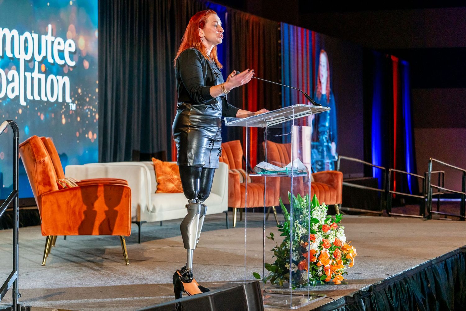 A person speaks on stage at a conference with orange chairs and blue lighting in the background.