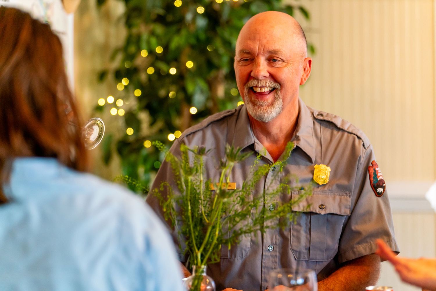 Park ranger in uniform shares a cheerful moment with visitor in front of Christmas tree lights.
