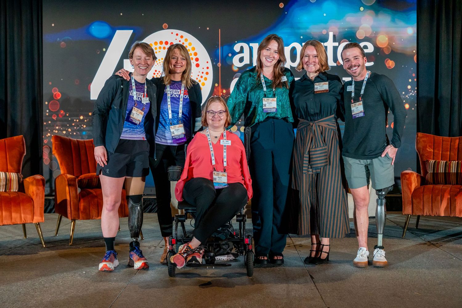 A group of conference attendees wearing lanyards pose together on stage at a tech event with blue lighting.