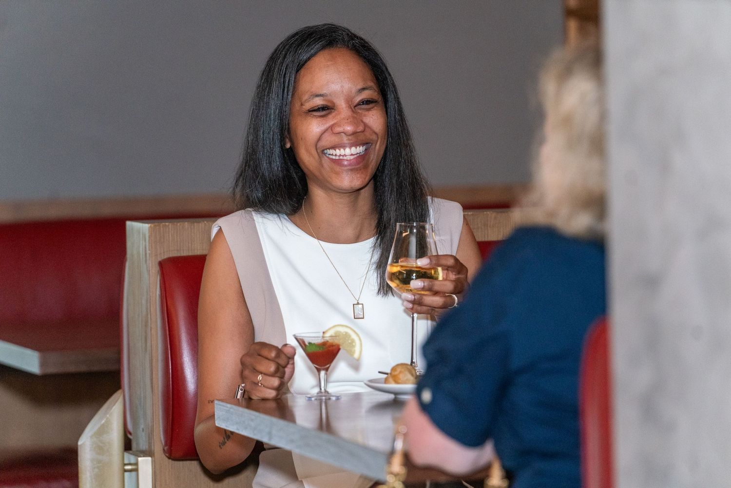 A smiling individual enjoying drinks at a restaurant table with wine glasses and comfortable booth seating.