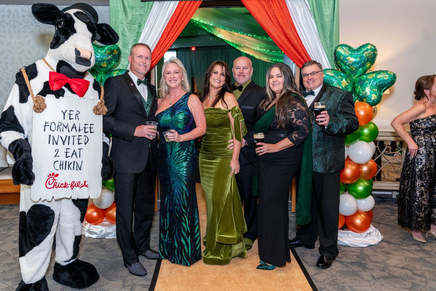 People in formal attire gather at a party with green and red decorations and balloons against a light-colored wall.