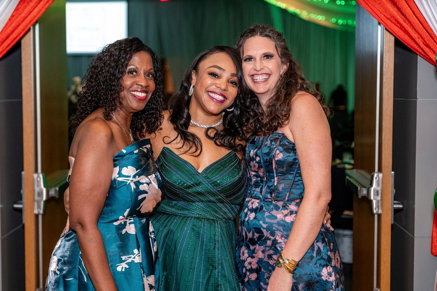 Three women in elegant floral and solid dresses pose together at a formal event with red and green decor.