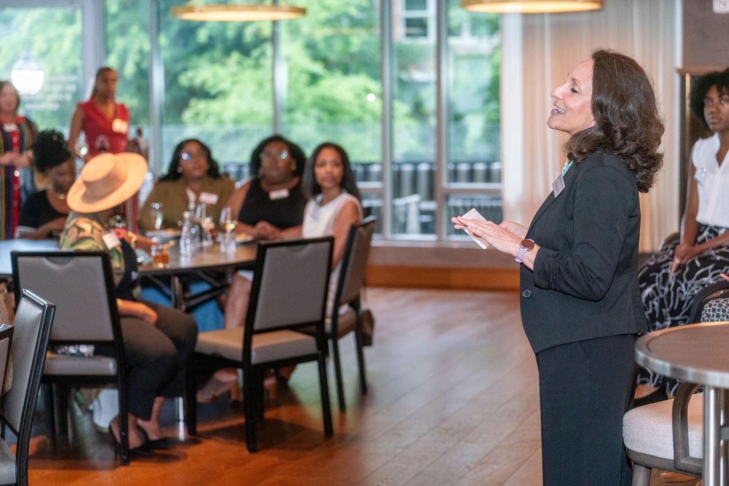 A speaker in business attire presents to an engaged audience in a modern restaurant setting with pendant lighting and windows.
