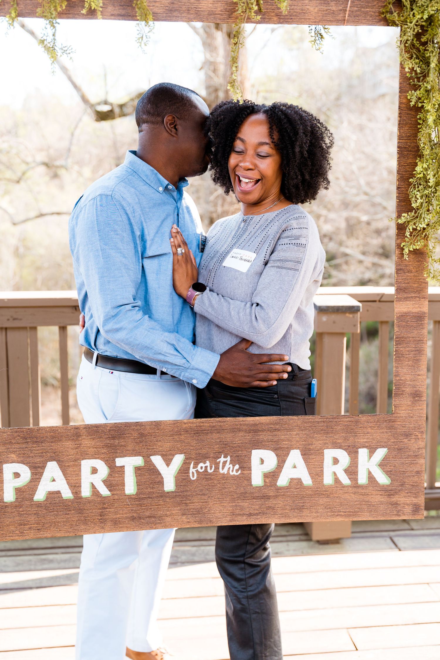 A joyful couple poses with a wooden Party for the Park sign frame outdoors on a deck.