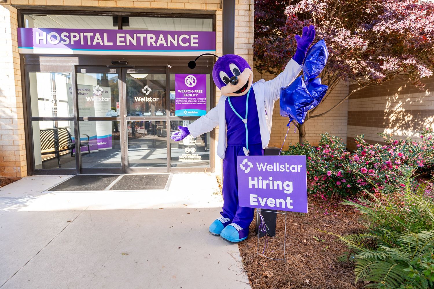 Hospital entrance decorated with blue mascots and a WellStar Rising Event sign displayed near landscaped garden.