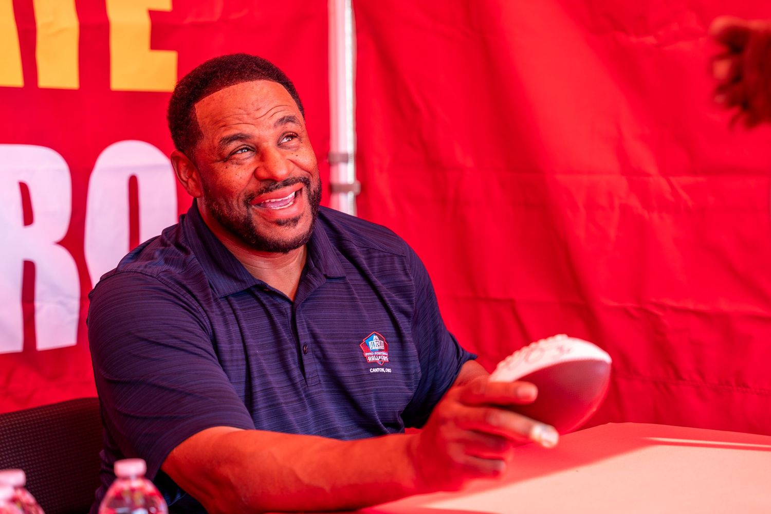 NBA player smiles while signing autographs at a promotional event with red backdrop.