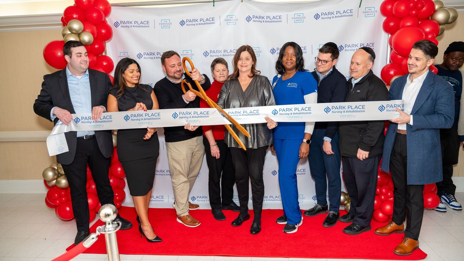 Group of people participate in ribbon cutting ceremony with red balloon decorations.