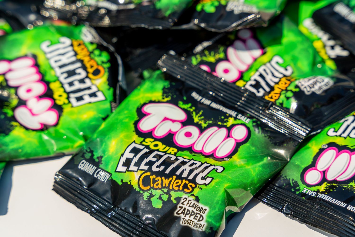 Several packages of Trolli Electric sour gummy candies in black and neon green wrappers scattered on a light surface.